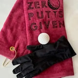 Red embroidered golf towel with 'Zero Putts Given,' golf ball, and black glove on white background.