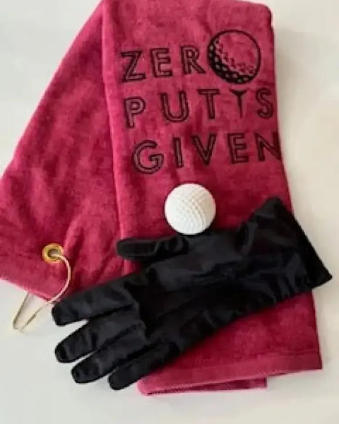 Red embroidered golf towel with 'Zero Putts Given,' golf ball, and black glove on white background.